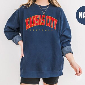 May include: Navy blue sweatshirt with the text "KANSAS CITY FOOTBALL" in red and yellow lettering. The sweatshirt has long sleeves with rolled-up cuffs. A navy blue circle with the word "NAVY" is in the upper right corner.