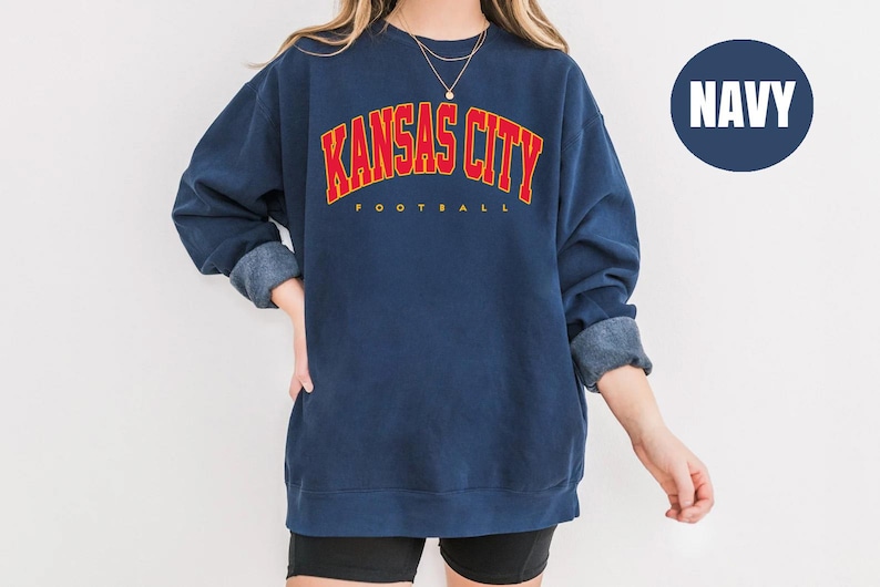 May include: Navy blue sweatshirt with the text "KANSAS CITY FOOTBALL" in red and yellow lettering. The sweatshirt has long sleeves with rolled-up cuffs. A navy blue circle with the word "NAVY" is in the upper right corner.