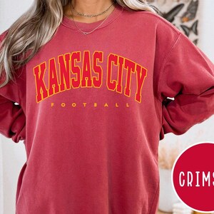 May include: Crimson-colored sweatshirt with the text "KANSAS CITY FOOTBALL" in yellow block letters. The sweatshirt is a solid color with a crew neck. A red circle with the word "CRIMSON" in white is in the lower right corner.