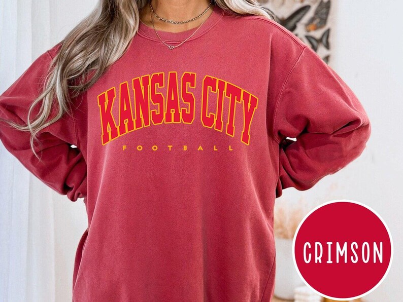 May include: Crimson-colored sweatshirt with the text "KANSAS CITY FOOTBALL" in yellow block letters. The sweatshirt is a solid color with a crew neck. A red circle with the word "CRIMSON" in white is in the lower right corner.