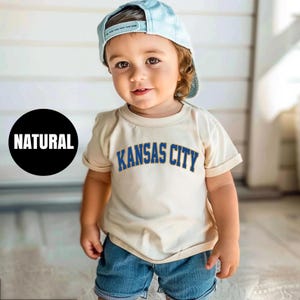 Peut inclure: Un jeune enfant portant une casquette de baseball bleu clair et un t-shirt cr&egrave;me avec l'inscription "KANSAS CITY" en bleu. Le t-shirt est associ&eacute; &agrave; un short en jean. Un cercle noir avec le mot "NATURAL" est &agrave; gauche.