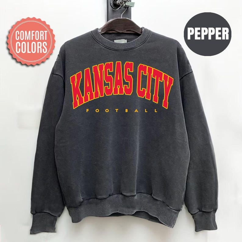 May include: A dark gray sweatshirt with the words "KANSAS CITY FOOTBALL" in red and yellow lettering. The sweatshirt has a crew neck and long sleeves. The words "Comfort Colors" are in a pink circle on the left side, and the word "PEPPER" is in a black circle on the right.