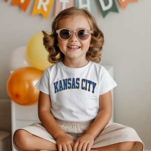 Peut inclure: Une jeune enfant portant des lunettes de soleil et un t-shirt blanc avec l'inscription "KANSAS CITY" en bleu. L'enfant sourit et est assis les jambes crois&eacute;es. Des ballons et une banni&egrave;re sont en arri&egrave;re-plan.