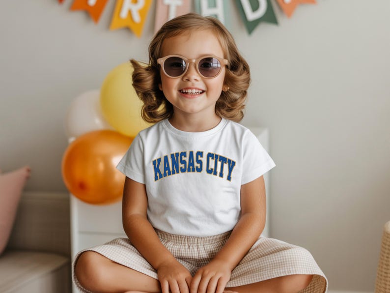 Peut inclure: Une jeune enfant portant des lunettes de soleil et un t-shirt blanc avec l'inscription "KANSAS CITY" en bleu. L'enfant sourit et est assis les jambes crois&eacute;es. Des ballons et une banni&egrave;re sont en arri&egrave;re-plan.