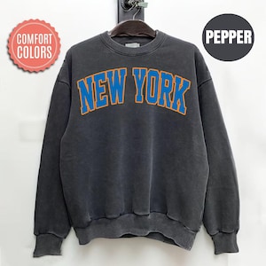 May include: A dark gray sweatshirt with the text "NEW YORK" in blue letters with orange outlines. The sweatshirt has a crew neck and long sleeves. The image also includes the text "COMFORT COLORS" and "PEPPER" in a circle.