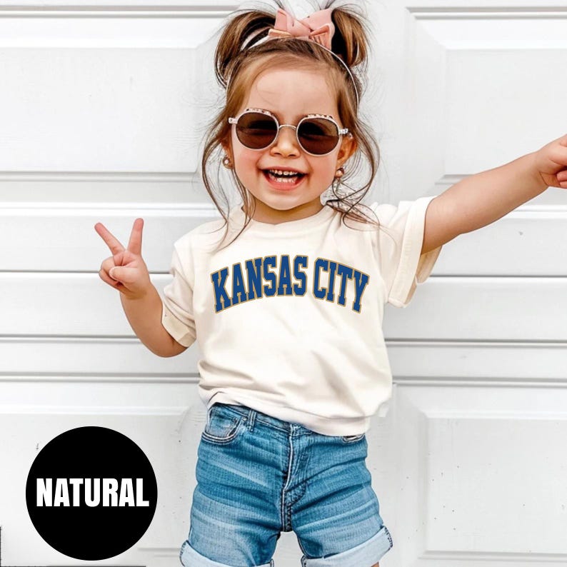 Peut inclure: Un enfant porte un t-shirt cr&egrave;me avec l'inscription "KANSAS CITY" en bleu. L'enfant porte &eacute;galement un short en jean et des lunettes de soleil. Un cercle noir avec le mot "NATURAL" est en bas &agrave; gauche.