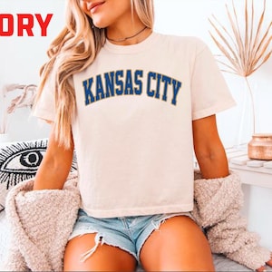 May include: Ivory-colored t-shirt with the text "KANSAS CITY" in blue lettering. The shirt is short-sleeved and appears to be made of a soft material. The word "IVORY" is in red in the top left corner.