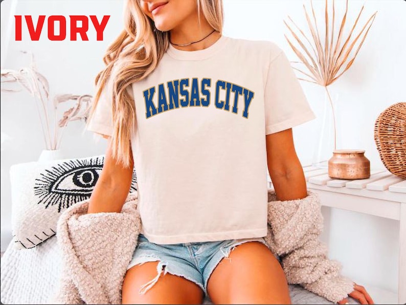 May include: Ivory-colored t-shirt with the text "KANSAS CITY" in blue lettering. The shirt is short-sleeved and appears to be made of a soft material. The word "IVORY" is in red in the top left corner.