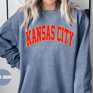 May include: Blue jean colored sweatshirt with the text "KANSAS CITY" in red arched lettering with a yellow outline. The word "FOOTBALL" is printed in yellow below the main text. The sweatshirt is a long-sleeved crew neck.