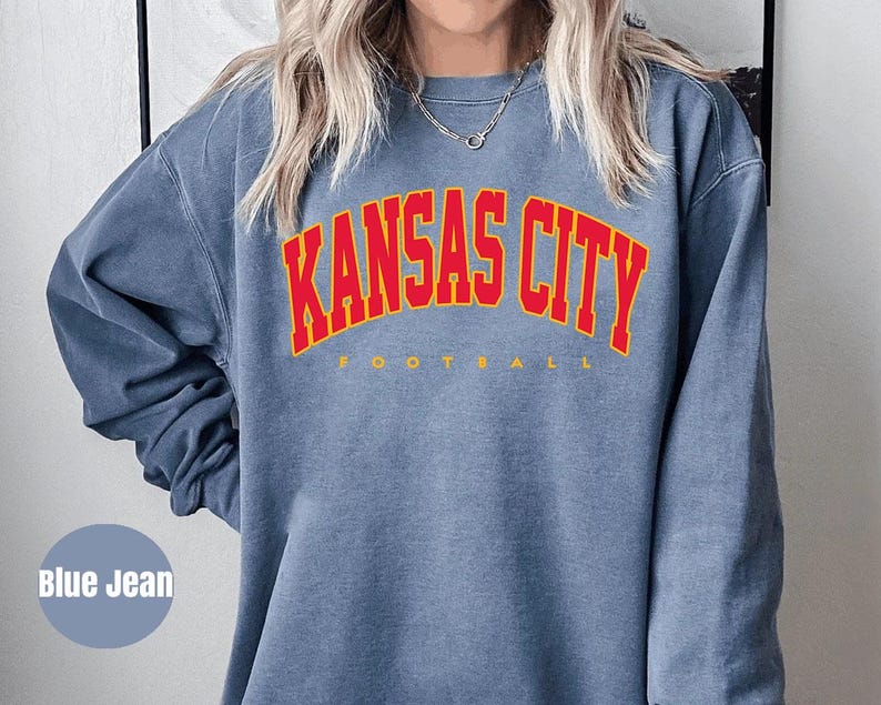 May include: Blue jean colored sweatshirt with the text "KANSAS CITY" in red arched lettering with a yellow outline. The word "FOOTBALL" is printed in yellow below the main text. The sweatshirt is a long-sleeved crew neck.