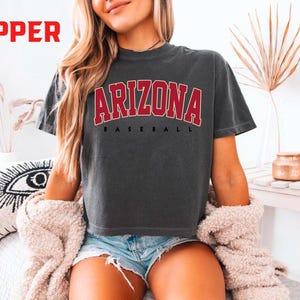 May include: Dark gray t-shirt with the word "ARIZONA" in red block letters arched across the chest, with "BASEBALL" below. The word "PEPPER" is in red in the upper left corner. The shirt is worn with denim shorts.