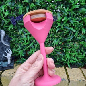 May include: A bright pink wine glass-shaped bottle stopper with a wooden top. The stopper is held in front of a green leafy background. The stopper has a wide base and a long stem, and the top is a light brown color.