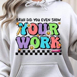 Op de afbeelding: Een lichtgrijze hoodie met de tekst "BRUH DID YOU EVEN SHOW YOUR WORK" in kleurrijke, gestikte letters. Het ontwerp bevat madeliefjes, sterren en potloden, tegen een zwart-witte geblokte rand.