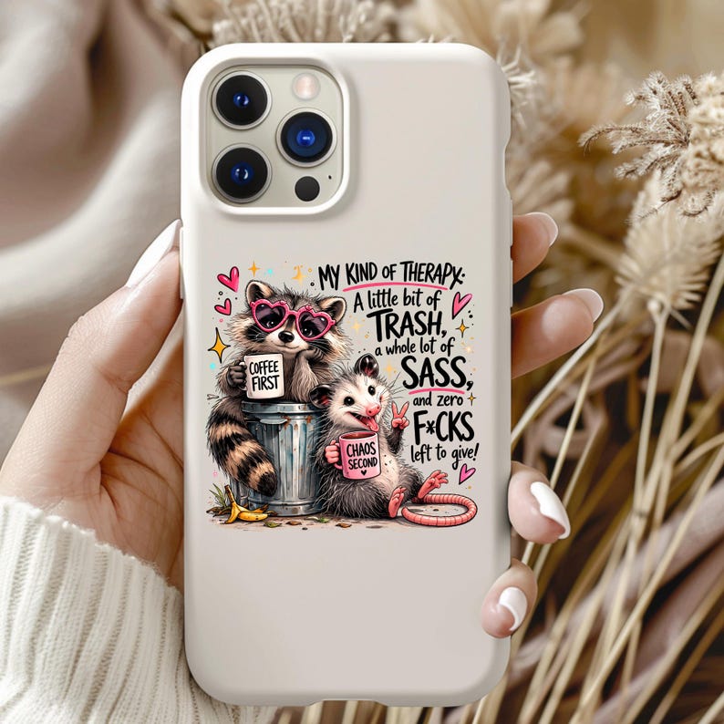May include: A white phone case featuring a cartoon raccoon wearing pink heart-shaped sunglasses and a possum. The raccoon holds a mug that says "COFFEE FIRST", and the possum holds a mug that says "CHAOS SECOND". Text on the case reads "MY KIND OF THERAPY: A little bit of TRASH, a whole lot of SASS, and zero F*CKS left to give!"