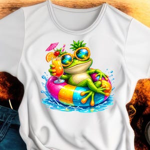 May include: White t-shirt featuring a cartoon frog wearing sunglasses and a pineapple on its head, floating in a colorful inflatable ring. The frog is holding a cocktail with a cherry and orange slice. The design is surrounded by water splashes.