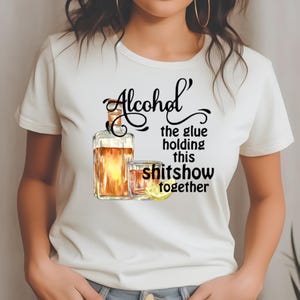 May include: White t-shirt featuring a watercolor illustration of a liquor bottle and a glass of amber-colored liquid with a lemon slice. The text reads "Alcohol the glue holding this shitshow together."