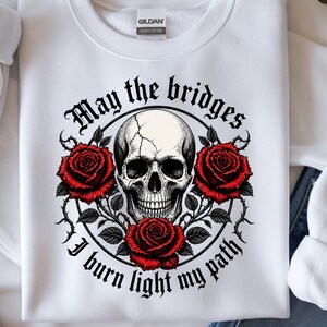 May include: White sweatshirt with a graphic design featuring a skull, red roses, and the text "May the bridges I burn light my path." The design is centered on the chest. The sweatshirt has a crew neck and long sleeves.