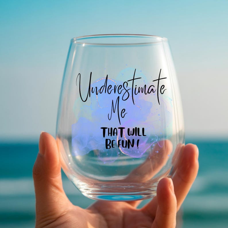Może przedstawiać: Przezroczysty kieliszek do wina bez n&oacute;żki z napisem "Underestimate Me That Will Be Fun!" czarnym pismem. Kieliszek ma niebieski i fioletowy wz&oacute;r akwarelowy. Kieliszek jest trzymany na tle niebieskiego oceanu.
