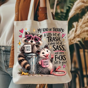 May include: Off-white tote bag featuring a raccoon wearing heart-shaped sunglasses and an opossum. The raccoon holds a mug that says "COFFEE FIRST," and the opossum holds a mug that says "CHAOS SECOND." Text on the bag reads "MY KIND OF THERAPY."