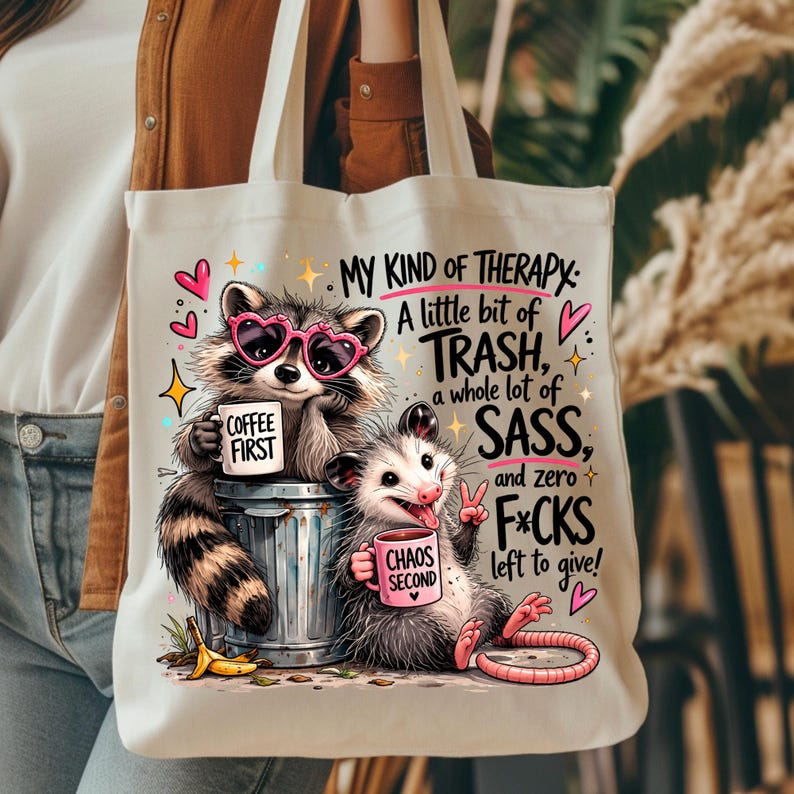 May include: Off-white tote bag featuring a raccoon wearing heart-shaped sunglasses and an opossum. The raccoon holds a mug that says "COFFEE FIRST," and the opossum holds a mug that says "CHAOS SECOND." Text on the bag reads "MY KIND OF THERAPY."