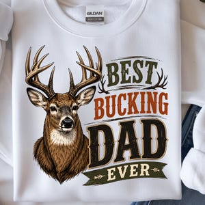 May include: White sweatshirt with a deer graphic and the text "BEST BUCKING DAD EVER" in green, brown, and black. The deer has large antlers and a detailed brown and tan coat. The sweatshirt is made of heavy cotton.