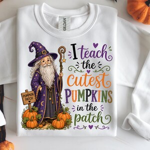 Può includere: Felpa bianca con un mago dei cartoni animati in abito e cappello viola, con un bastone, zucche e la scritta "I teach the cutest pumpkins in the patch". Perfetta per Halloween.