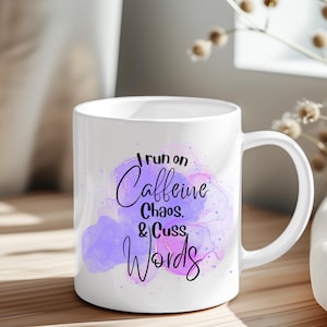 Może przedstawiać: Biały ceramiczny kubek z uchwytem. Kubek zawiera napis "I run on Caffeine, Chaos, & Cuss Words" czarną czcionką na tle akwarelowego wzoru w odcieniach fioletu i r&oacute;żu. Kubek stoi na drewnianej powierzchni.