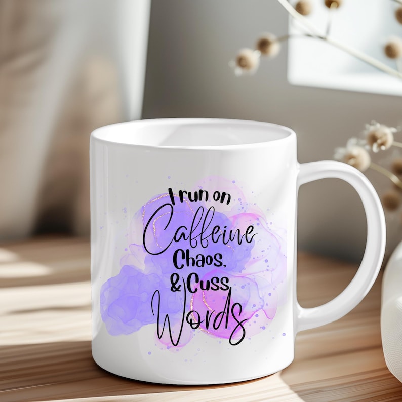 Może przedstawiać: Biały ceramiczny kubek z uchwytem. Kubek zawiera napis "I run on Caffeine, Chaos, & Cuss Words" czarną czcionką na tle akwarelowego wzoru w odcieniach fioletu i r&oacute;żu. Kubek stoi na drewnianej powierzchni.
