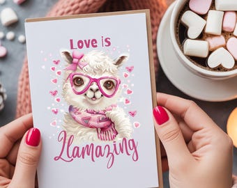 May include: A greeting card with a cartoon llama wearing pink glasses, a bow, and a scarf. The card reads "Love is Llamazing" in pink script. The card is held in hands with red nail polish, with a cup of hot chocolate and marshmallows in the background.