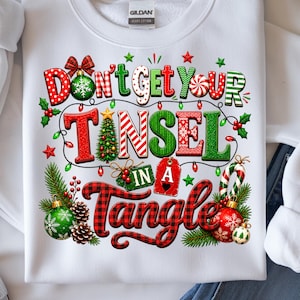 May include: A white sweatshirt featuring a colorful Christmas-themed graphic with the text "DON'T GET YOUR TINSEL IN A TANGLE." The design includes red and green lettering, a small Christmas tree, ornaments, pine branches, holly, and string lights.