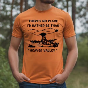 Könnte beinhalten: Orangefarbenes T-Shirt mit schwarzer Grafik und Text. Der Text lautet "THERE'S NO PLACE I'D RATHER BE THAN "BEAVER VALLEY"" über einer stilisierten Illustration einer Atompilzwolke und Landschaft.
