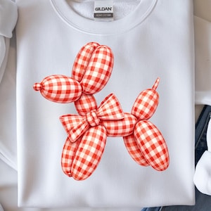 May include: White sweatshirt featuring a red and white gingham balloon dog design. The balloon dog has a bow tie and is made of several balloon shapes. The sweatshirt has a crew neck and long sleeves.