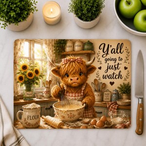 May include: A rectangular cutting board featuring a fluffy Highland cow wearing a red and white checkered apron, whisking batter in a bowl. The text reads Y'all going to just watch. The scene includes a flour jar, rolling pin, eggs, and sunflowers in a kitchen setting.