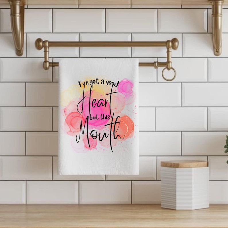 Może przedstawiać: Biały ręcznik kuchenny z napisem "I've got a good Heart but this Mouth" czcionką. Ręcznik ma kolorowe plamy w stylu akwareli w kolorze r&oacute;żowym, ż&oacute;łtym i pomarańczowym. Ręcznik wisi na złotym wieszaku na ręczniki.