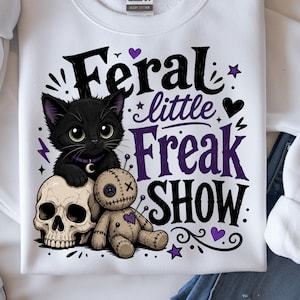 May include: White sweatshirt featuring a black cat, skull, and voodoo doll graphic. The text reads "Feral little Freak Show" in black and purple. The design includes stars, hearts, and lightning bolts.