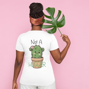 Op de afbeelding: Een wit T-shirt met korte mouwen voorzien van een afbeelding van een groene cactus in een bruine pot met een droevige uitdrukking, een kleine roze bloem en de tekst "Not A HUGGER" in zwarte en groene letters op de achterkant.