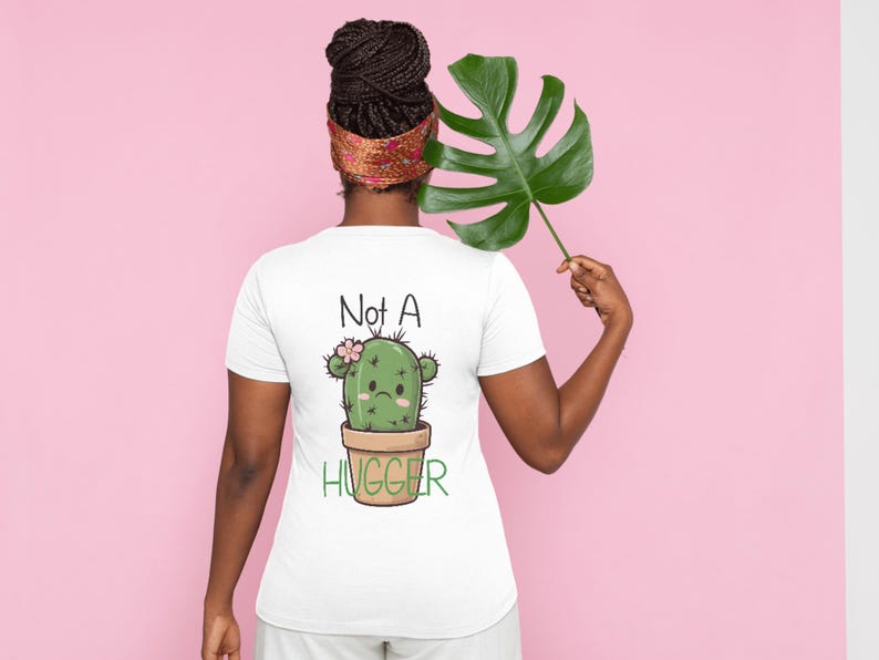Op de afbeelding: Een wit T-shirt met korte mouwen voorzien van een afbeelding van een groene cactus in een bruine pot met een droevige uitdrukking, een kleine roze bloem en de tekst "Not A HUGGER" in zwarte en groene letters op de achterkant.