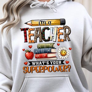 Puede incluir: Una sudadera con capucha gris claro con un diseño gráfico colorido. El diseño incluye un lápiz, las palabras "I'm a TEACHER", libros, una manzana, una margarita con una cara sonriente y la frase "WHAT'S YOUR SUPERPOWER?" en naranja.