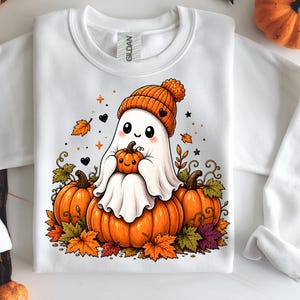 May include: White sweatshirt with a cartoon ghost wearing an orange knitted hat and holding a small pumpkin. The ghost is sat on three orange pumpkins, surrounded by autumn leaves and decorative elements.