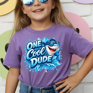 Op de afbeelding: Een lila T-shirt met een cartoon haai met een zonnebril en een pet. De tekst "ONE Cool DUDE" is blauw, met een spetterontwerp. Het shirt heeft korte mouwen.