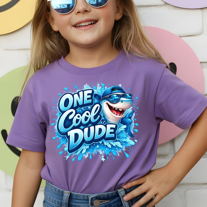 Op de afbeelding: Een lila T-shirt met een cartoon haai met een zonnebril en een pet. De tekst "ONE Cool DUDE" is blauw, met een spetterontwerp. Het shirt heeft korte mouwen.