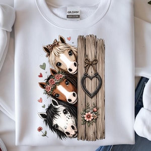 Puede incluir: Sudadera blanca con un diseño de tres caballos asomándose por detrás de una valla de madera. Los caballos tienen crines de diferentes colores y están adornados con coronas de flores y lazos. Una decoración metálica en forma de corazón cuelga de la valla.