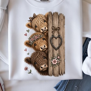 May include: White sweatshirt featuring a graphic of three Highland cows peeking from behind a wooden fence. The cows have brown fur and are adorned with a bow, flower crown, and flowers. A metal heart hangs on the fence, with pink flowers at the bottom.