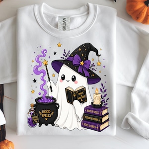 Puede incluir: Sudadera blanca con un fantasma de dibujos animados con un sombrero de bruja y leyendo un libro titulado "Magic Mischief". El fantasma sostiene una varita, con un caldero de "Good Spells" y libros apilados.
