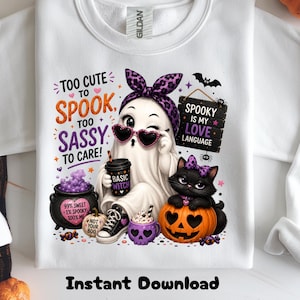May include: A white sweatshirt featuring a graphic of a ghost wearing a purple leopard print headband and heart-shaped sunglasses, holding a cup labeled Basic Witch. Beside it are a black cauldron, a pumpkin, and a black cat with a purple bow.