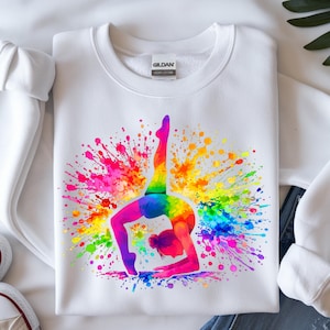 May include: White sweatshirt featuring a colorful watercolor design of a gymnast in a bridge pose. The gymnast is depicted in rainbow colors, with a vibrant splash of paint in the background. The sweatshirt has a crew neck and long sleeves.