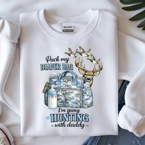 以下が含まれることがあります： おむつバッグ、哺乳瓶、鹿、飛んでいるアヒルが描かれたグラフィックデザインの白いスウェットシャツ。「おむつバッグを詰めて。パパと狩りに行く」と書かれています。