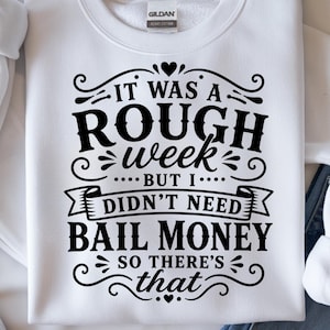 May include: White sweatshirt with the text "IT WAS A ROUGH week... BUT I... DIDN'T NEED BAIL MONEY SO THERE'S that" in black. The sweatshirt has a crew neck and the Gildan logo is visible.