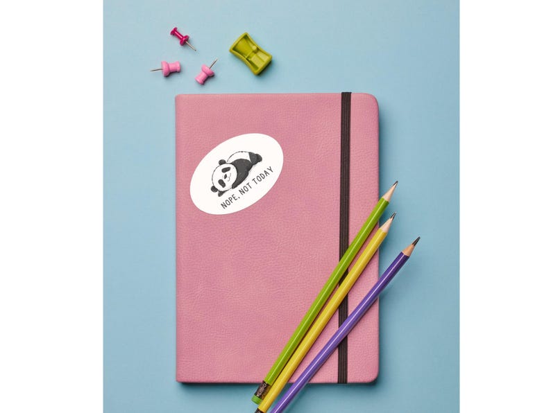 Op de afbeelding: Een roze notitieboek met een zwarte elastische band, voorzien van een ovale sticker van een liggende panda met de tekst "NOPE, NOT TODAY". Drie kleurpotloden in groen, geel en paars liggen op de kaft, samen met drie kleine punaises.