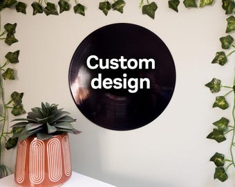 May include: A black vinyl record with the words "Custom design" in white lettering. The record is mounted on a light-colored wall, with artificial ivy and a potted succulent plant in the foreground.
